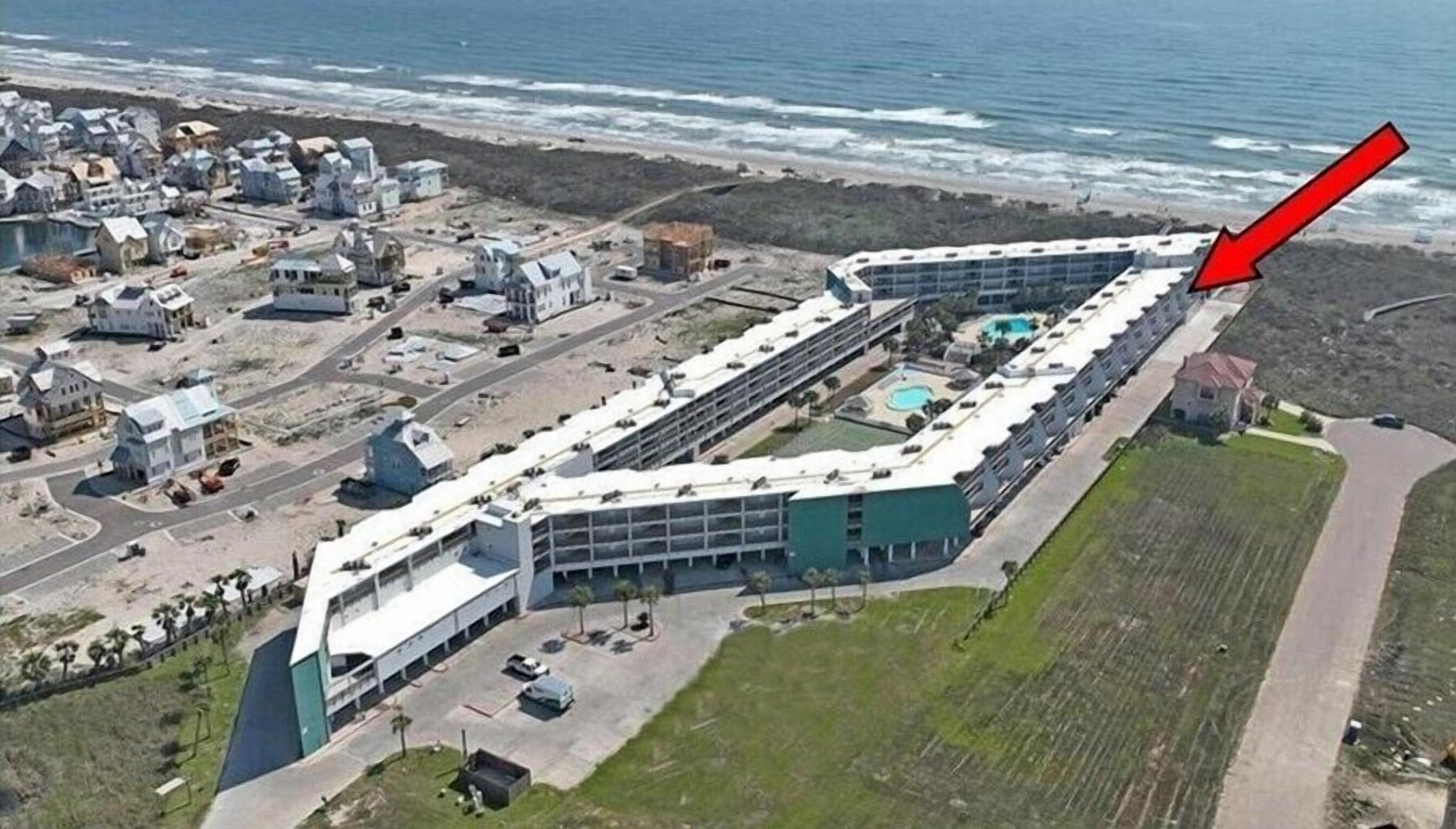 An aerial view of a coastal hotel with a long, curved building, a parking lot, and a beach in the background.