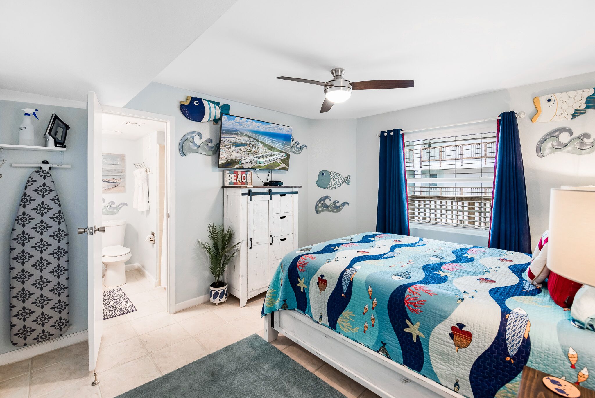 A bedroom with a bed, a ceiling fan, a window with curtains, a wall-mounted TV, and a bathroom visible through an open door.