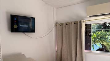 Um quarto com uma cama de solteiro, televisão na parede, janela com cortinas e aparelho de ar condicionado.