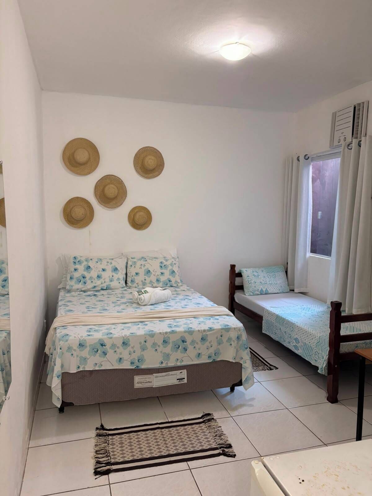 A bedroom with a bed, a bench, a window with curtains, and wall-mounted hats.