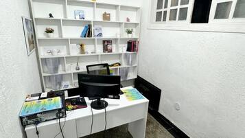 Um quarto pequeno com paredes brancas, uma escrivaninha, estantes e uma janela.