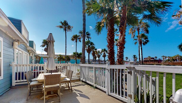 A covered patio with a table and chairs, surrounded by a white fence, palm trees, and a clear blue sky. - Charming Townhouse,  Shared pool, Great Location (Port Aransas)