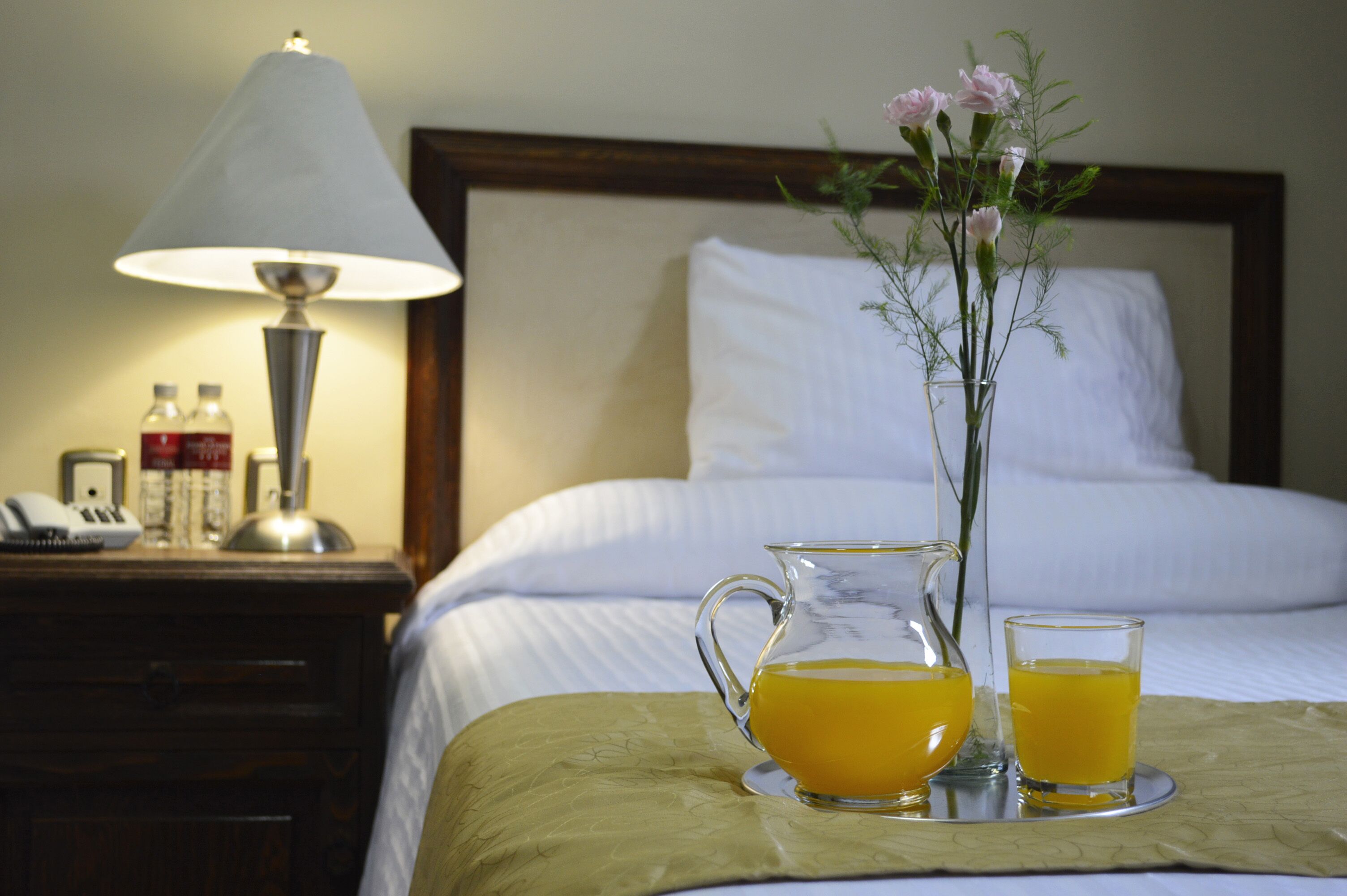 Una cama bien hecha con ropa blanca, una mesita de noche con una lámpara, un jarrón con flores y una bandeja con un jarro de jugo de naranja y un vaso.