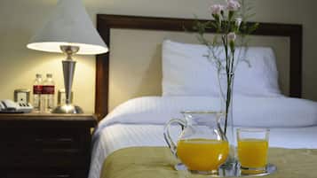 Una cama bien hecha con ropa blanca, una mesita de noche con una lámpara, un jarrón con flores y una bandeja con un jarro de jugo de naranja y un vaso.