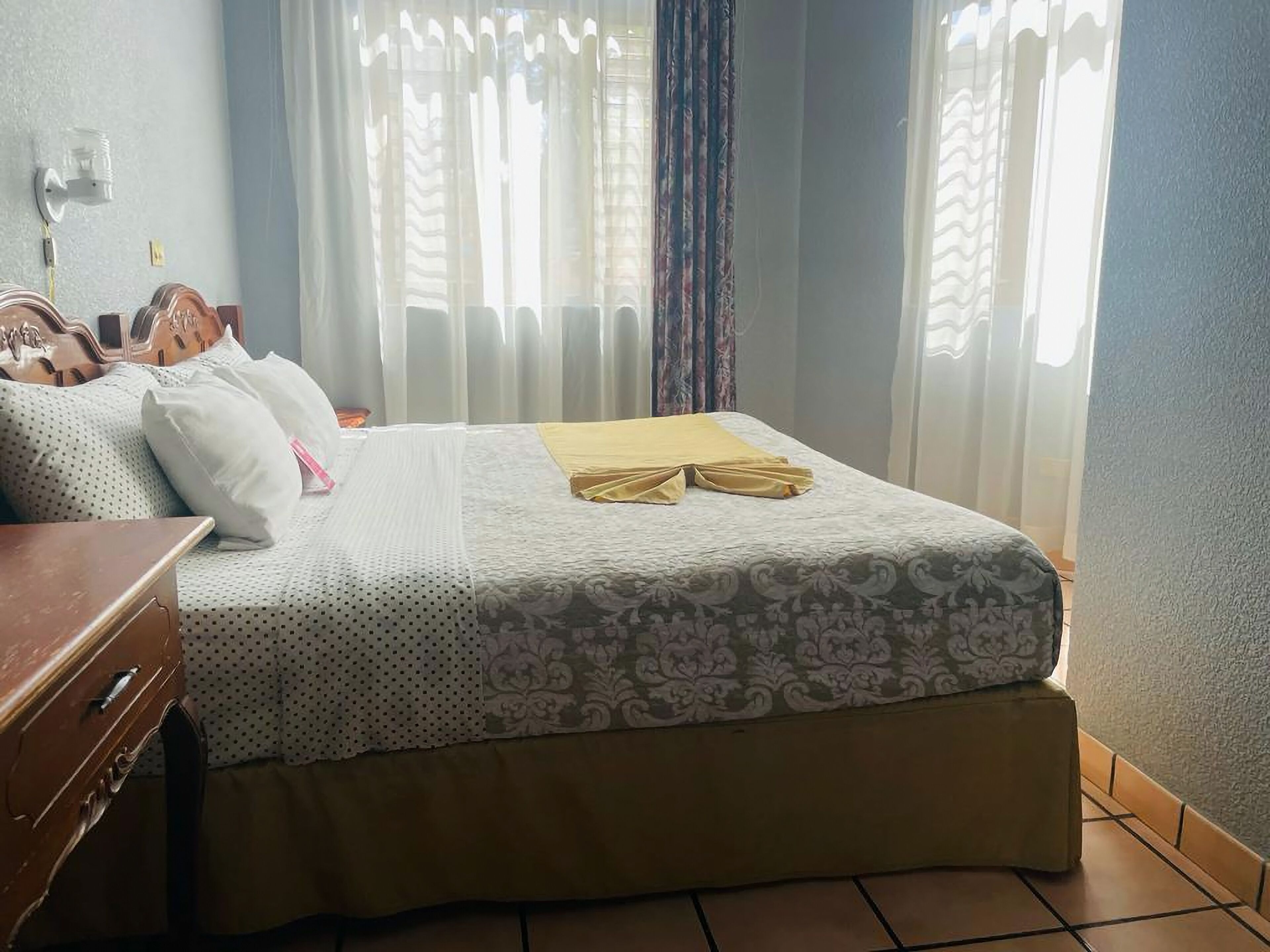 A neatly made bed with a patterned bedspread and two towels, a wooden nightstand, and a wall-mounted lamp.
