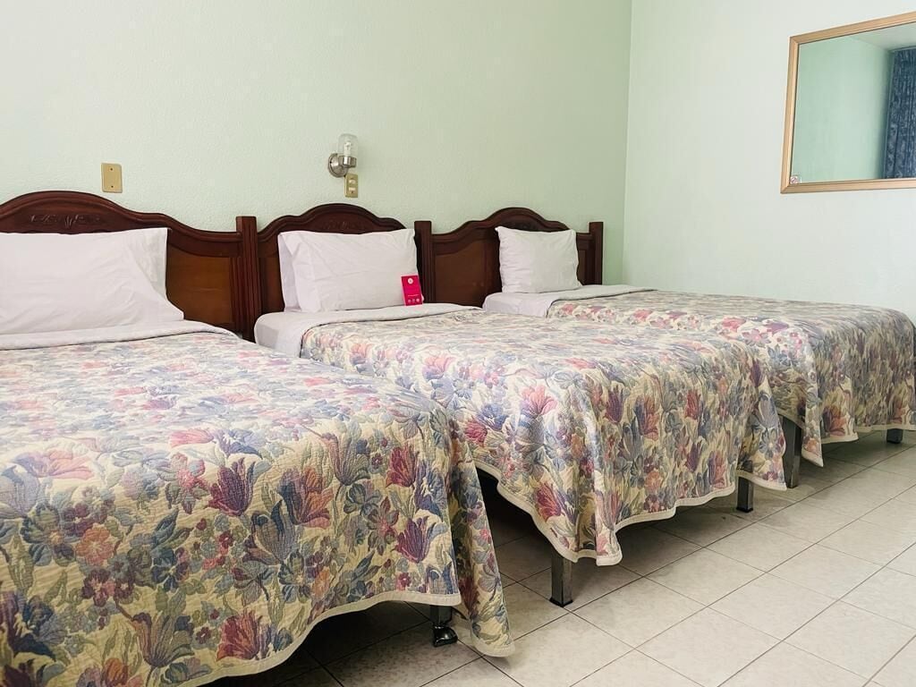 A hotel room with two single beds, each with a floral-patterned bedspread and wooden headboards. There is a nightstand with a lamp and a red object on it. The room has a tiled floor and a mirror on the wall.