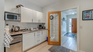 A modern kitchen with white cabinets, a microwave, and a coffee maker. There is a wooden door leading to a hallway with a rug. - The Cedar - Downtown, walking distance to all the shops and restaurants! (Banff)