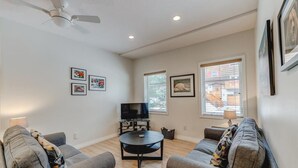 A living room with two grey sofas, a black coffee table, a ceiling fan, and a TV on a stand. There are framed pictures on the walls and a window with blinds. - The Cedar - Downtown, walking distance to all the shops and restaurants! (Banff)