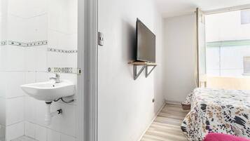 Habitación de hotel con pared de azulejos blancos, un televisor montado, una mesita de noche y una cama con cubrecama estampado.