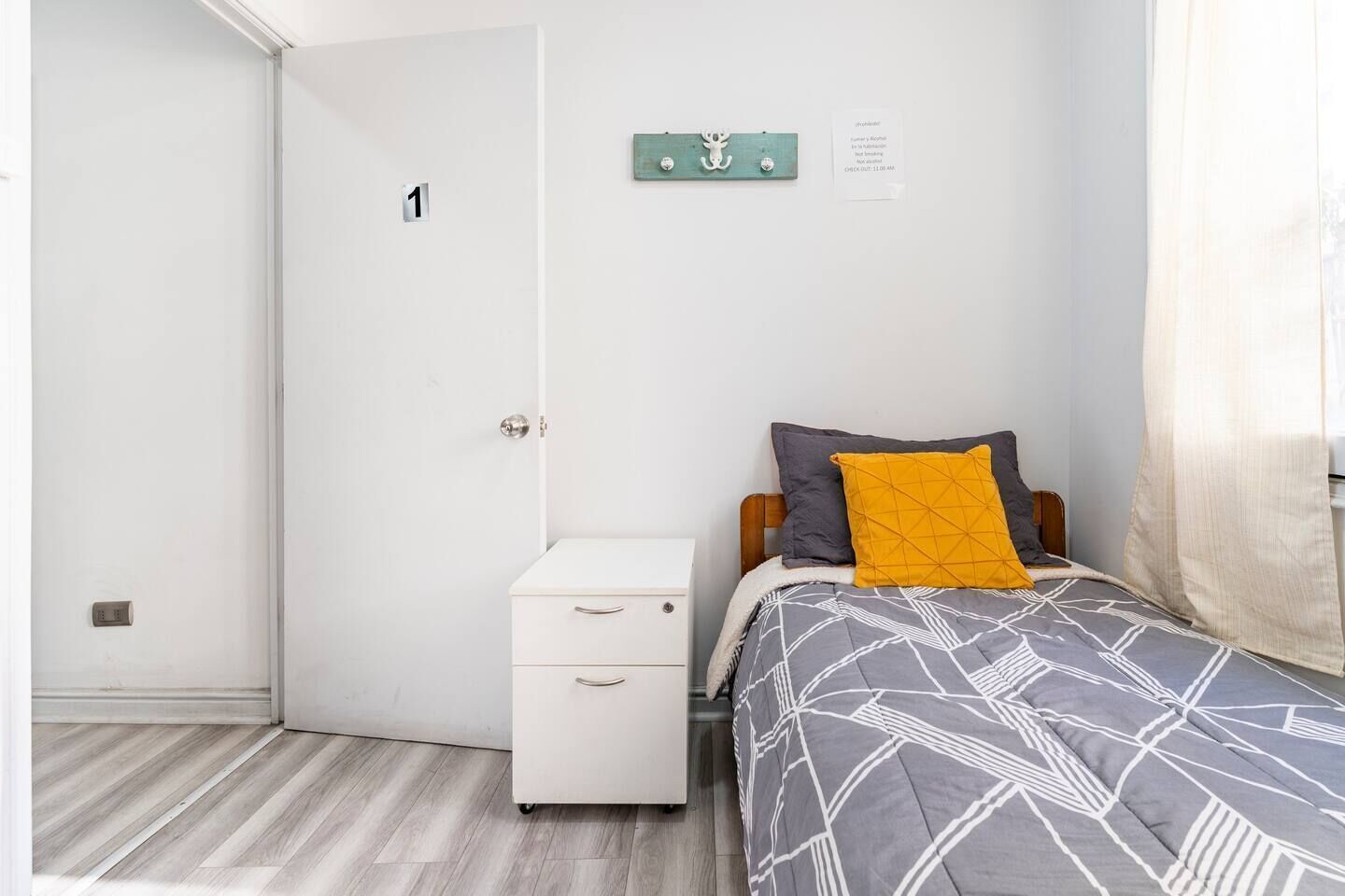 A bedroom with a bed, a nightstand, a wall-mounted light, and a door with a number.