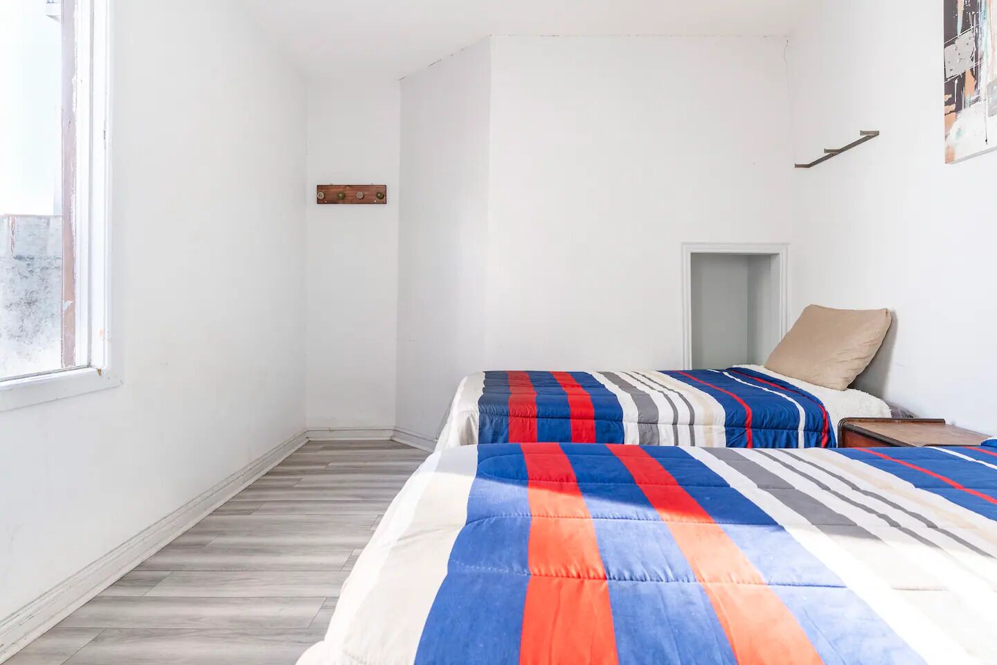 A small, bright room with two beds, a wooden floor, a window, and a door.