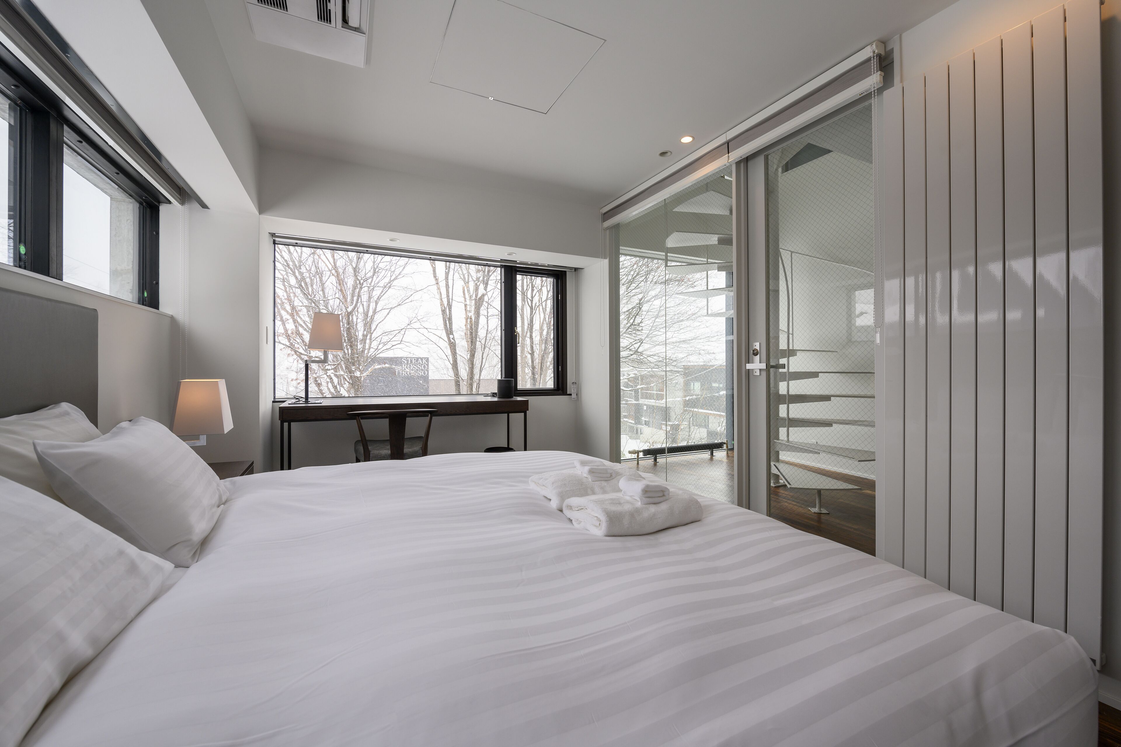 A modern bedroom with a large bed, a desk, and a sliding glass door leading to a balcony.