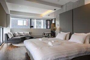 Una habitación de hotel moderna con una cama grande, un sofá, una mesita y vistas a la ciudad a través de amplios ventanales.