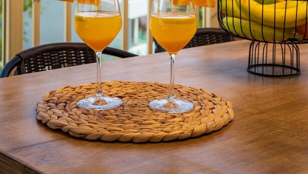 Zwei Gläser Orangensaft auf einer gewebten Tischmatte auf einem Holztisch, mit Blick auf das Meer und einer Topfpflanze im Hintergrund.
