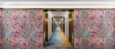 Ein Hotelgang mit floralem Wandpapier, einem blauen Teppich und einem langen Korridor, der zu einer Tür führt.