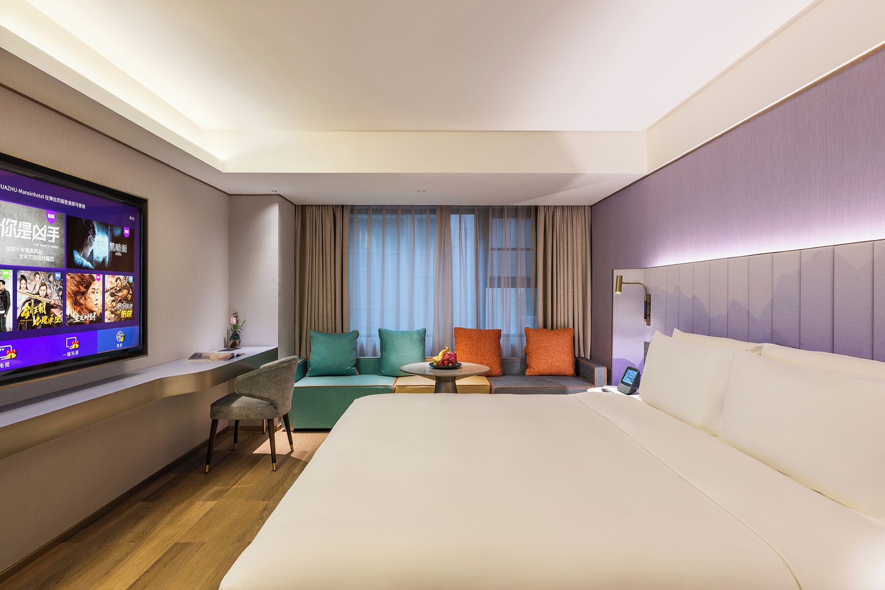 Habitación de hotel con cama, escritorio con computadora, televisor y zona de estar con cojines.