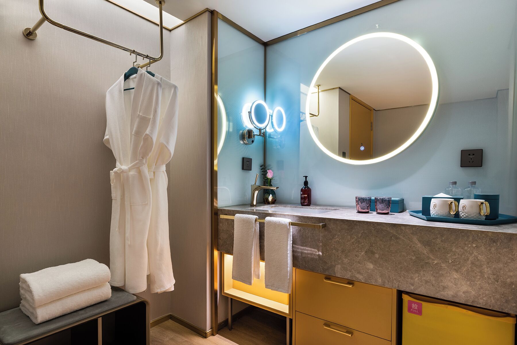 A modern bathroom with a large mirror, a robe hanging on a rack, a towel rack, and a vanity with a sink and toiletries.