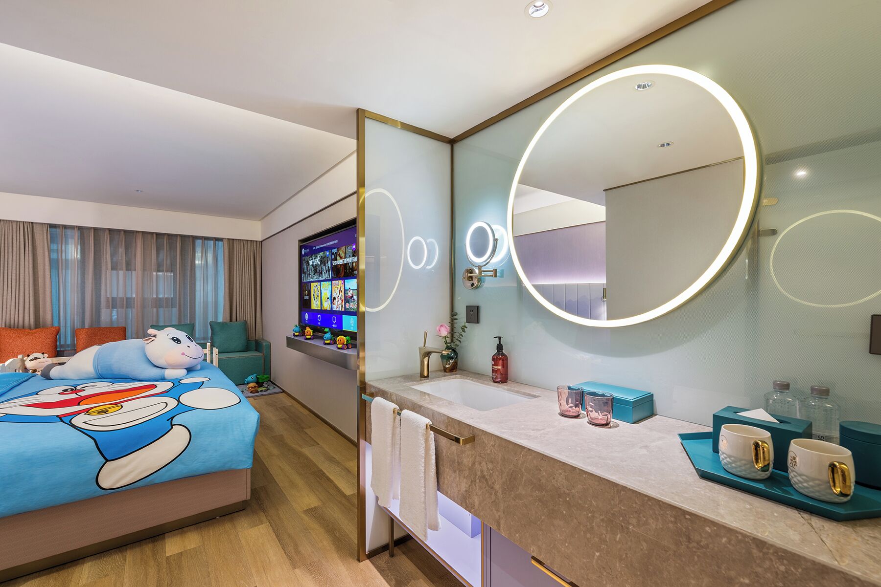 A modern hotel room with a large bed featuring a cartoon character design, a flat-screen TV, a vanity with a mirror, and a bathroom counter with toiletries.