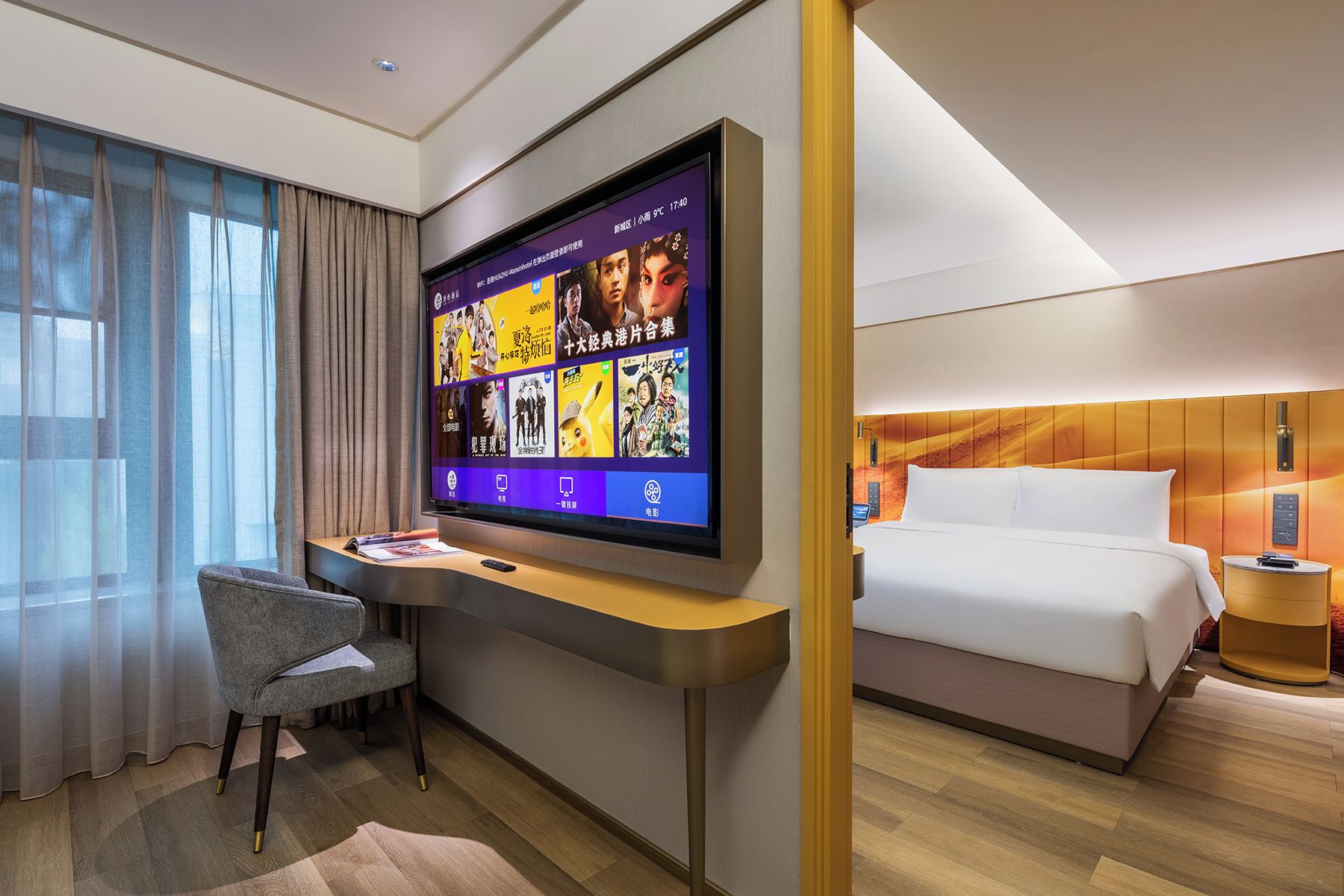 Ett hotellrum med en platt-TV som visar filmplakat, ett skrivbord med en stol, en säng med vita sängkläder och ett trägolv.