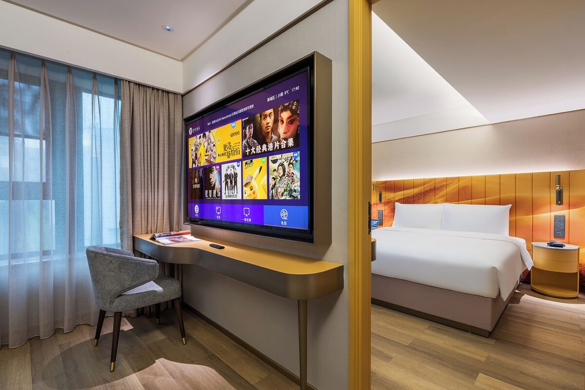 A hotel room with a flat-screen TV displaying movie posters, a desk with a chair, a bed with white bedding, and a wooden floor.