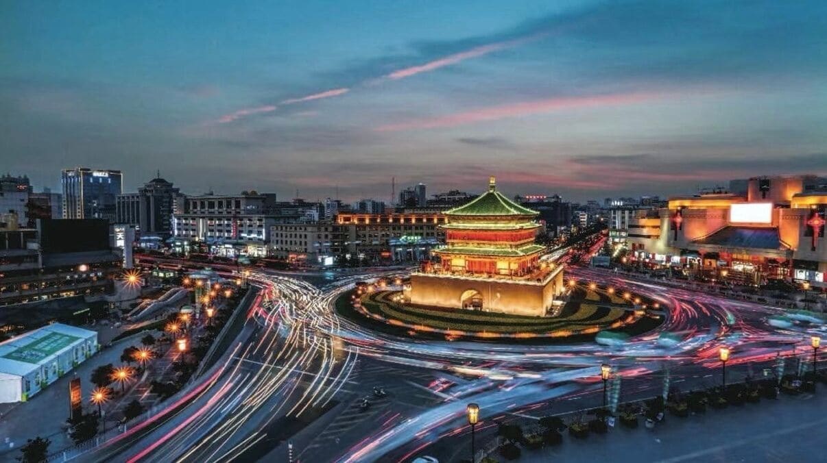 A cityscape at dusk with a prominent traditional building with a green roof and a circular plaza.