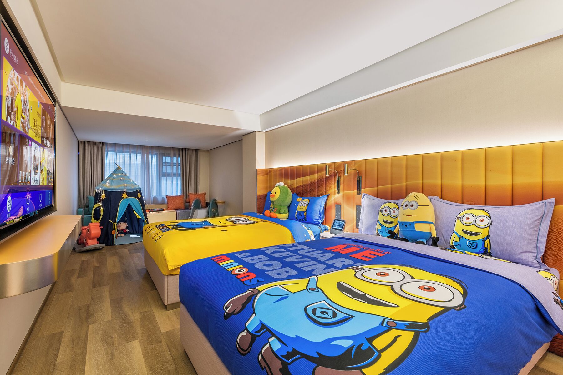 A hotel room with a large bed featuring Minion-themed bedding, a separate seating area with a tent-like structure, and a television on the left side.