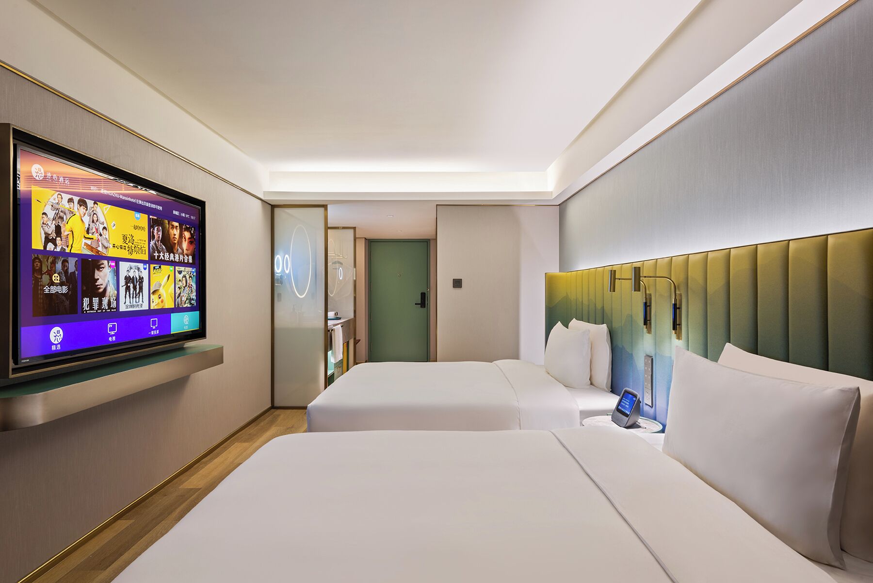 Ein Hotelzimmer mit großem Flachbildfernseher an der Wand, zwei Betten mit weißer Bettwäsche, einem modernen Kopfteil und einem kleinen Nachttisch mit Lampe.