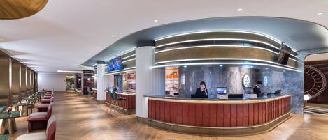 A modern hotel lobby with a curved reception desk, seating area with tables and chairs, and a bar.