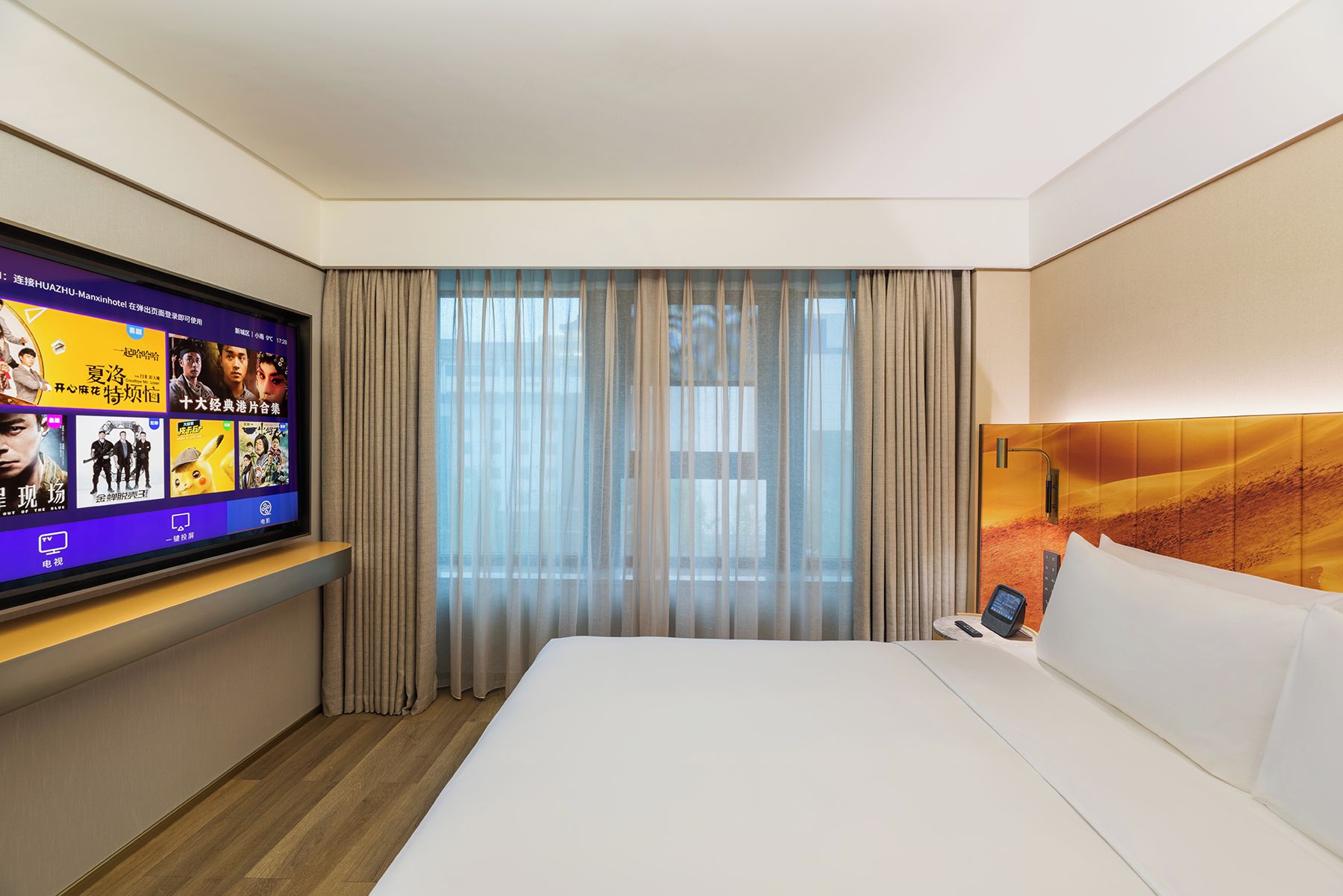 Habitación de hotel con cama, televisión en la pared, ventana con cortinas y cabecera con lámpara.