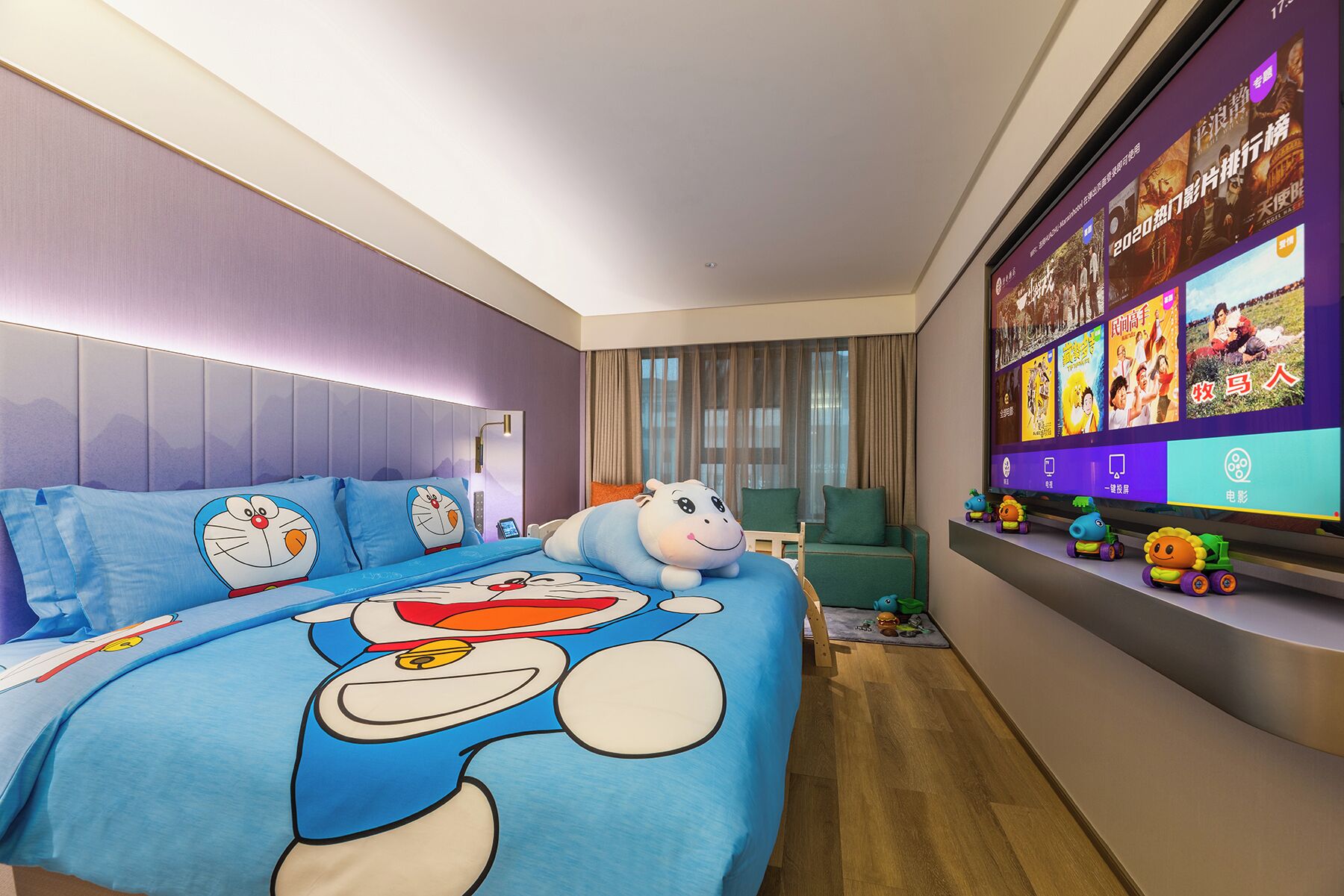 Ein Hotelzimmer mit einem großen Bett, das mit Bettwäsche mit Zeichentrickfiguren, einem Fernseher mit Filmplakaten und einem kleinen Regal mit Spielzeug ausgestattet ist.