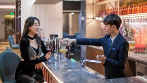 A bar area with a person pouring a drink into a glass. There are shelves with bottles and glasses in the background.