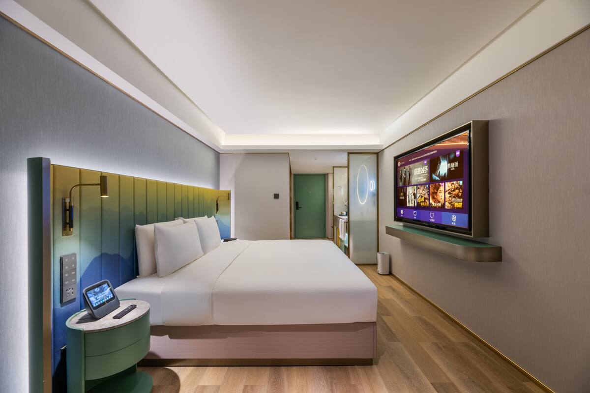 Una habitación de hotel moderna con una cama grande, una mesita de noche con una tableta, un televisor montado en la pared y una pared de acento verde.