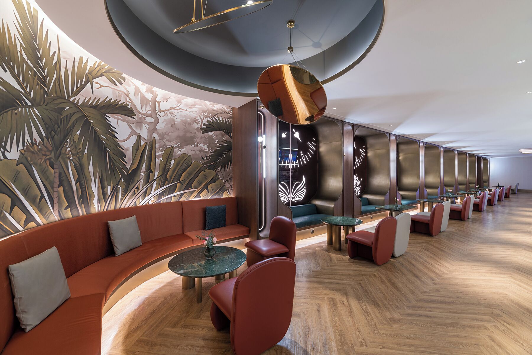A modern hotel lobby with a curved seating area, a mural of tropical foliage, and a unique hanging light fixture.