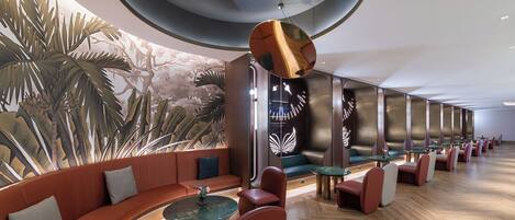 A modern hotel lobby with a curved seating area, a mural of tropical foliage, and a unique hanging light fixture.
