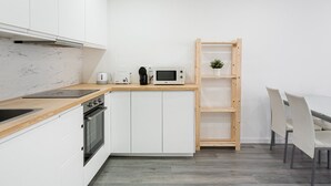Eine moderne Küche mit weißen Schränken, einer Holz Arbeitsplatte, einem eingebauten Ofen, einer Mikrowelle, einem Wasserkocher und einer Kaffeemaschine. Es gibt ein Holzregal und einen Esstisch mit Stühlen.