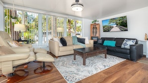 Ein Wohnzimmer mit beiger Couch, schwarzer Couch und Couchtisch aus Holz. Ein großes Fenster mit vertikalen Jalousien, eine Kristallleuchter und ein gerahmtes Bild an der Wand sind ebenfalls zu sehen.