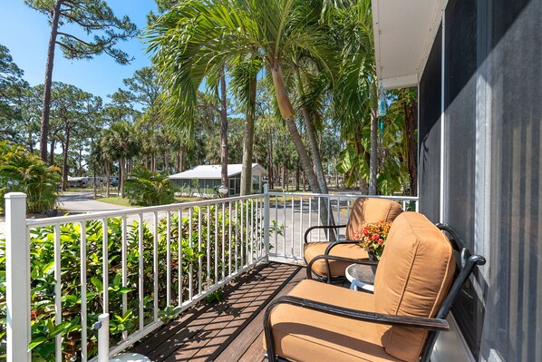 A balcony with two chairs and a table, surrounded by a white railing and lush greenery, including palm trees and various plants. - Cozy park home amenities galore! Near Legacy Bike trail & only 3 miles to beach. (Nokomis)