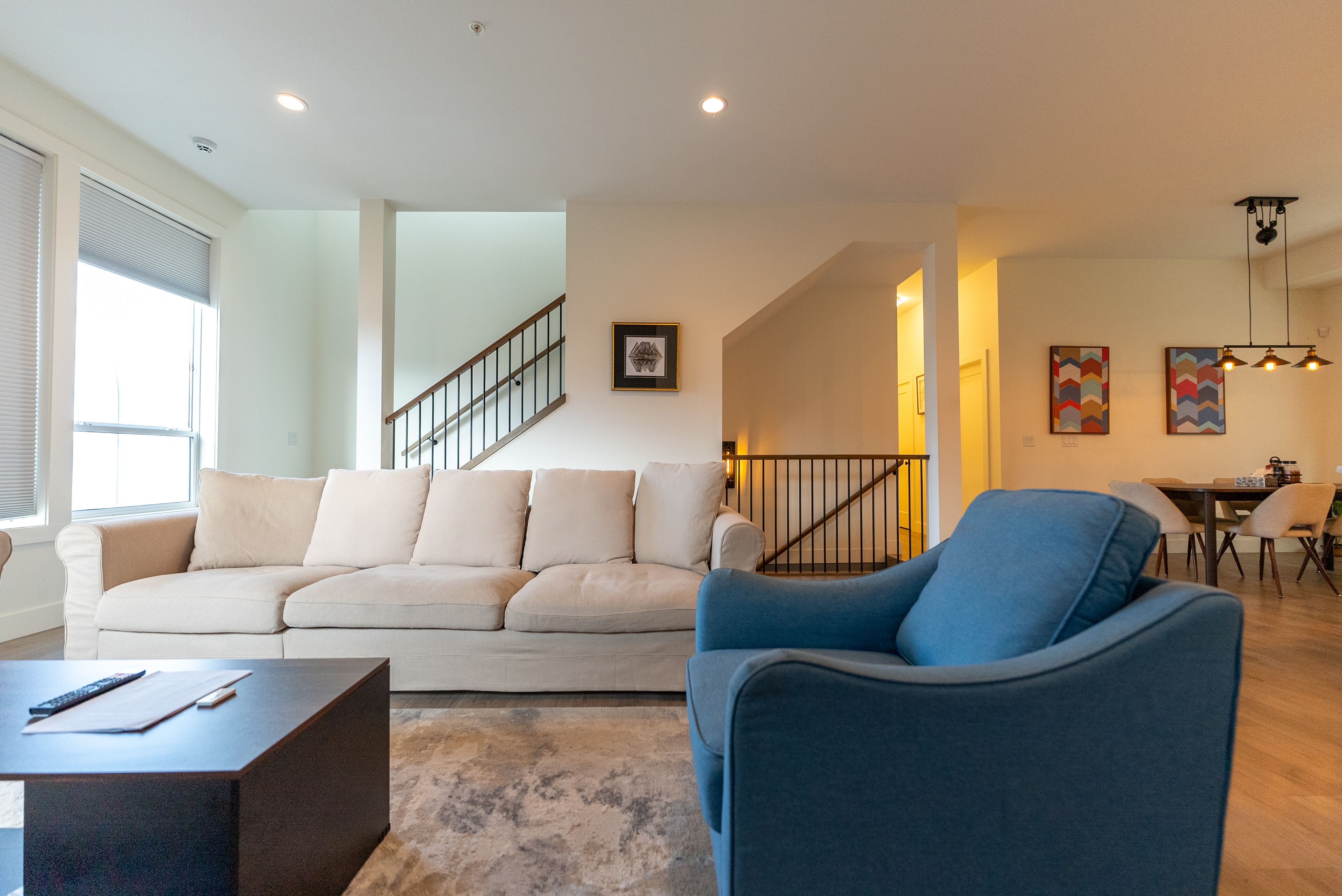 A modern living room with a beige sofa, a blue armchair, and a coffee table. There is a staircase leading to an upper level, and a dining area with a table and chairs.