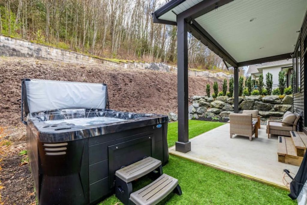 A hot tub with steps, a covered patio area with outdoor furniture, and a landscaped garden with a stone wall and trees.