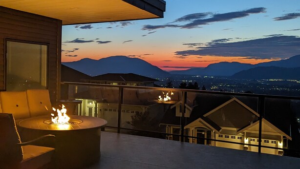 A balcony with a fire pit and seating, overlooking a cityscape and mountains at dusk. - A Perfect Mountain Retreat w/ Views, Hot Tub, Home Theatre, Games, PS5 & AC (Chilliwack)