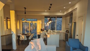 A modern living room with a sofa, a coffee table, and a blue armchair. There is a kitchen area with a refrigerator and a dining table with chairs. - A Perfect Mountain Retreat w/ Views, Hot Tub, Home Theatre, Games, PS5 & AC (Chilliwack)