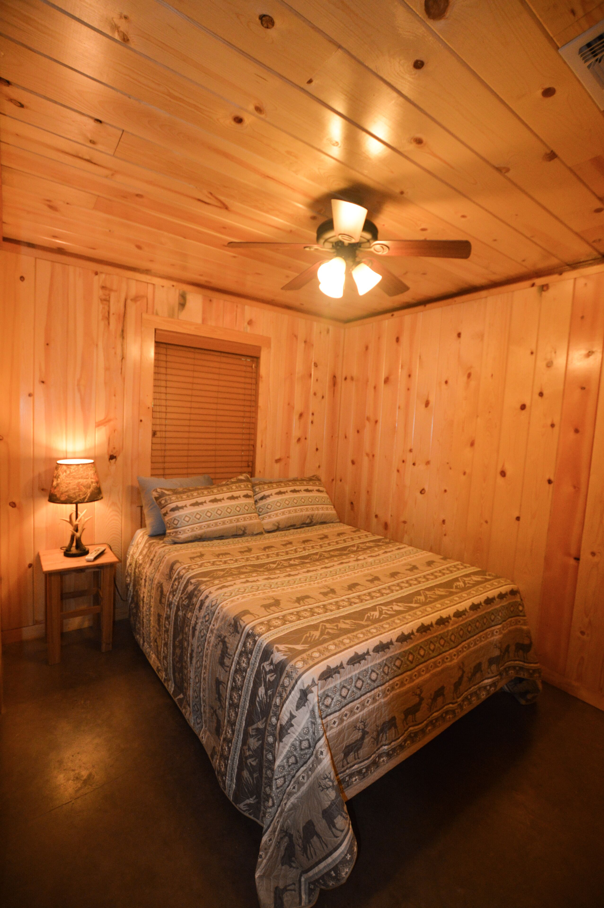 Um quarto com painéis de madeira, uma cama, uma mesa de cabeceira, um abajur e um ventilador de teto.