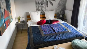 A bedroom with a bed, a nightstand, a lamp, a window with curtains, and a wall with a palm tree mural.