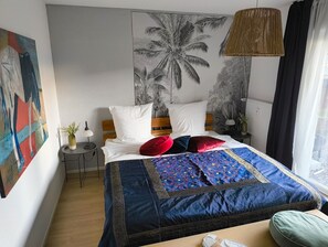 A bedroom with a bed, a nightstand, a lamp, a window with curtains, and a wall with a palm tree mural. - Apartment 'Haus Auryn' with Private Terrace, Shared Garden and Wi-Fi (Radolfzell am Bodensee)