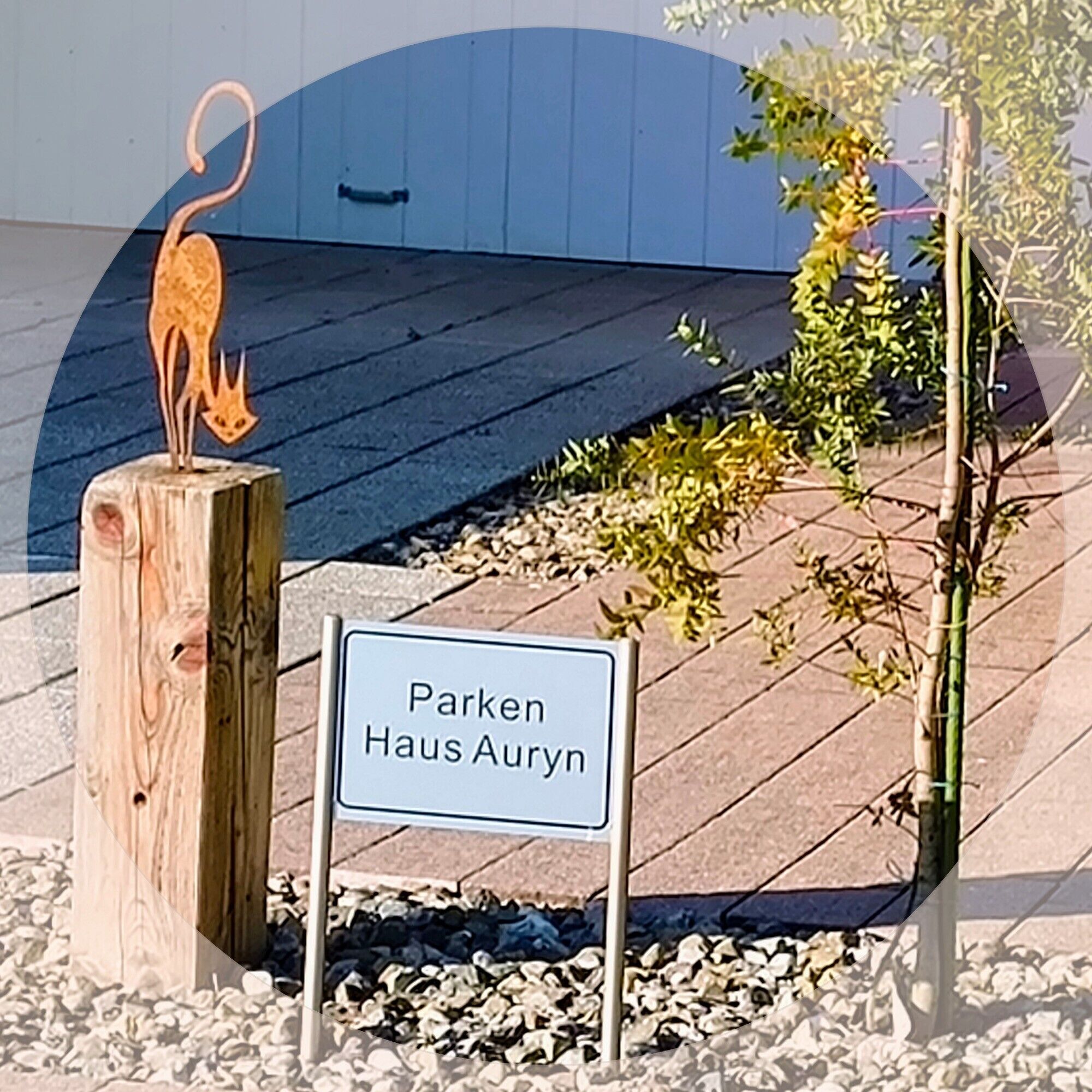 Un poteau en bois surmonté d’une sculpture de chat, une pancarte indiquant « Parken Haus Auryn », et une petite plante.