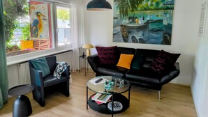 A living room with a black sofa, a glass coffee table, a painting, and a window with a view of greenery.