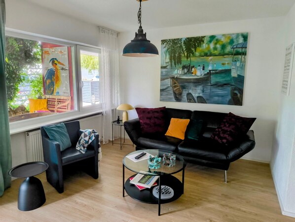 A living room with a black sofa, a glass coffee table, a painting, and a window with a view of greenery.