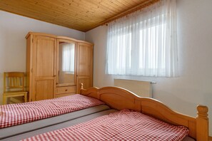 A bedroom with two single beds, a wooden wardrobe, a chair, and a window with sheer curtains.