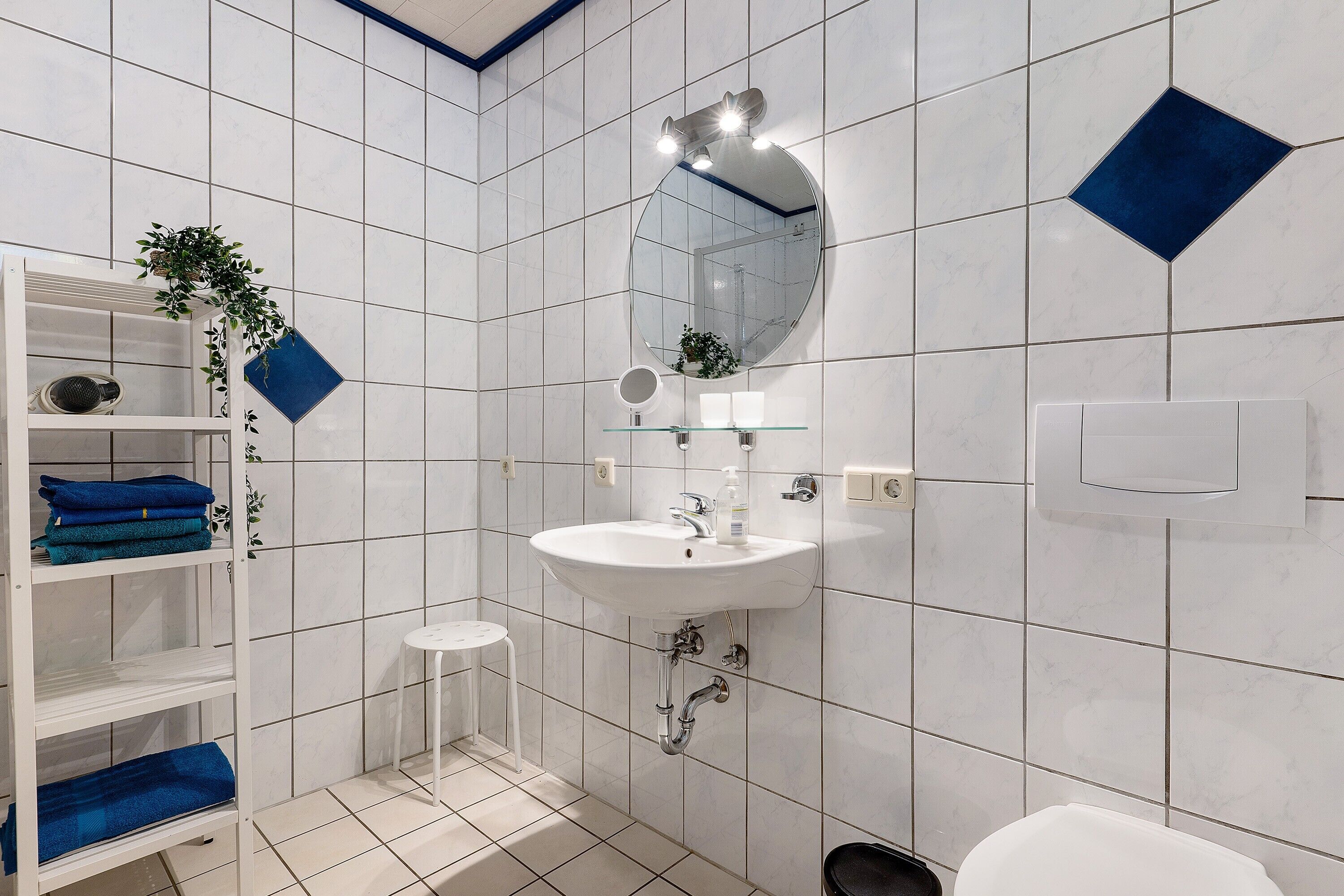 A bathroom with white tiled walls, a white sink, a mirror, a small stool, and shelves with folded towels.