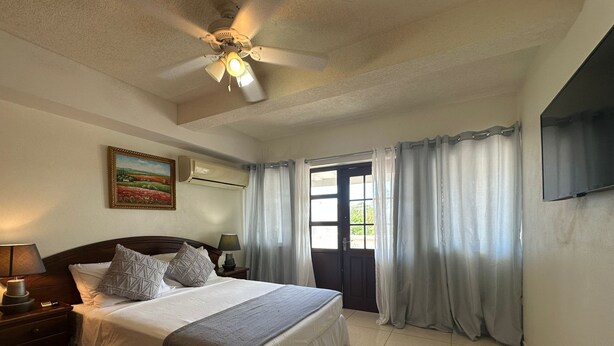 A hotel room with a bed, a ceiling fan, a painting, a TV, and a door with curtains. - St. Lawrence Suites - Suite Three (Oistins)